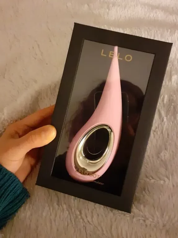 Ręka trzyma minimalistyczne, czarne opakowanie, w którym widać jasnoróżowy stymulator łechtaczki LELO Dot.